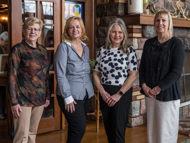 Four women in business stand indoors before wooden doors and a stone fireplace, smiling at the camera. Dressed in business casual attire, these female business leaders appear to be at a social or professional gathering.
