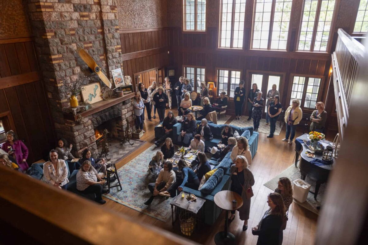 A group of female entrepreneurs gather in a spacious, rustic living room with high ceilings, wooden walls, a stone fireplace, large windows, and comfortable seating, engaging in lively conversation and activities.