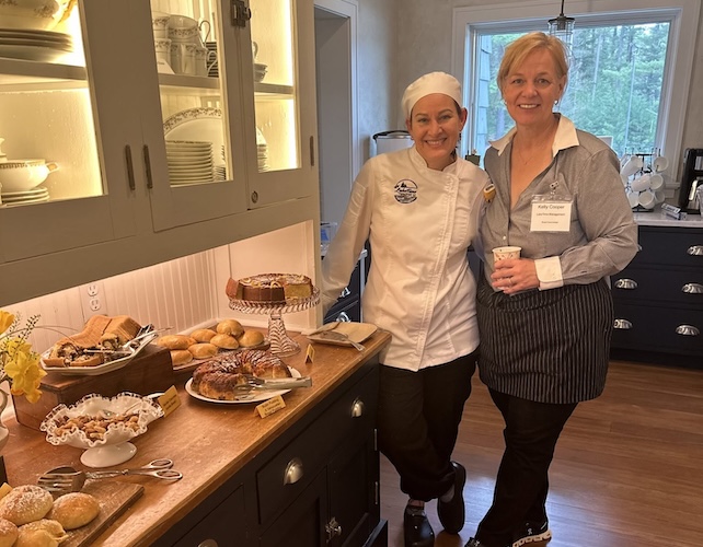 Two women entrepreneurs stand smiling in a cozy kitchen beside a counter filled with pastries and baked goods. One wears a chef’s uniform and hat; the other, a striped apron, holds a cup. Plates and cups are visible on the shelves behind them.