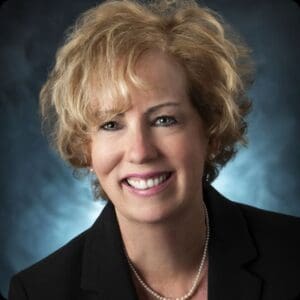 Smiling Executive Director Kathy Schmitz, with short, curly blond hair, wearing a black blazer and pearl necklace, is posed in front of a dark blue background representing VCEDC.